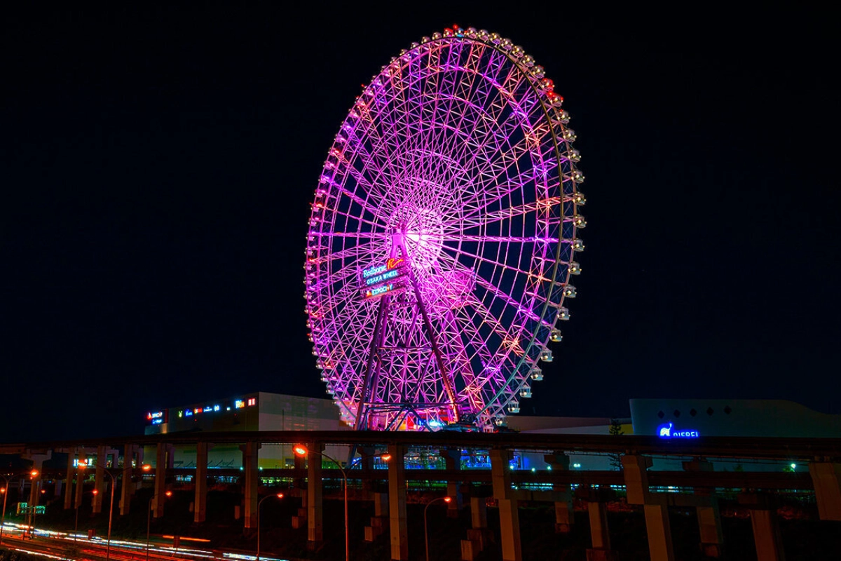 OSAKA WHEEL是一座巨大的夜景摩天輪，夜晚亮起紫色、粉色燈光，搭配現代購物中心、廣場燈光與親子旅客背影，一股溫馨感油然而生。